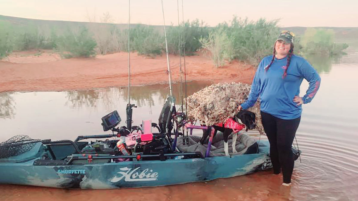 Erin de pie en aguas poco profundas con su perro Kaya y su kayak lleno de equipo de pesca
