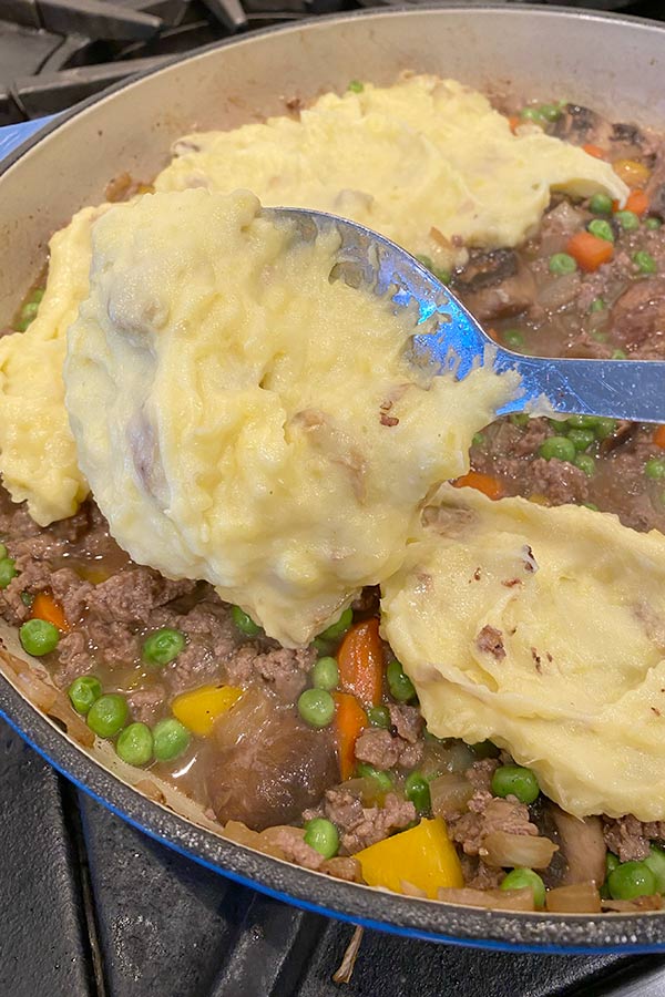 Mashed potatoes being added to elk meat, vegetables, and mushrooms in a saucepan