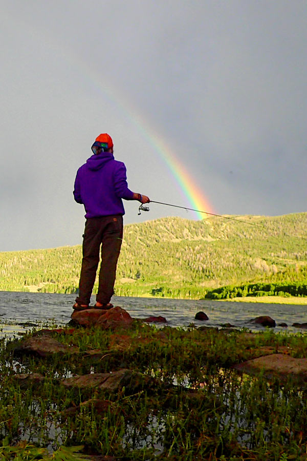 Man with a fishing pole, casting a line in a pond, under a cloudy sky with a rainbow