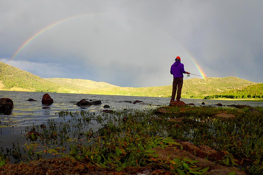 Hombre con una caña de pescar, lanzando una línea en un estanque, bajo un cielo nublado con un arco iris