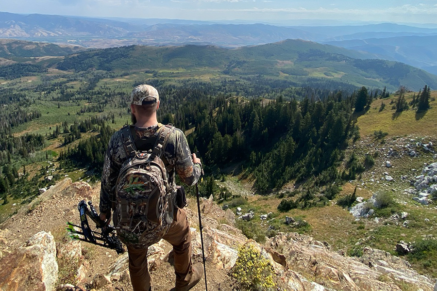 Bowhunter carrying a bow standing atop a mountain peak, overlooking a valley