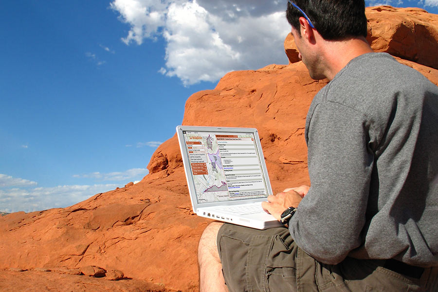 Man sitting near Utah&apos;s red rocks, browsing the Utah Hunt Planner on his laptop