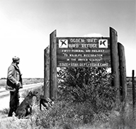 Waterfowl hunter, holding a rifle, with his dog at the entrance sign of the Ogden Bay Bird Refuge (now called the Ogden Bay Waterfowl Management Area)