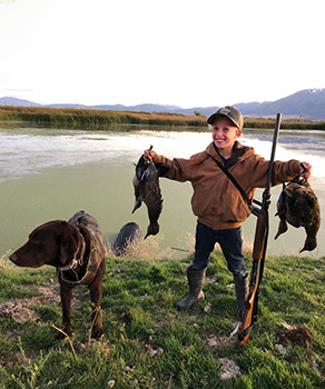 Young waterfowl hunter holding four harvested ducks, standing next to a hunting dog at a lake