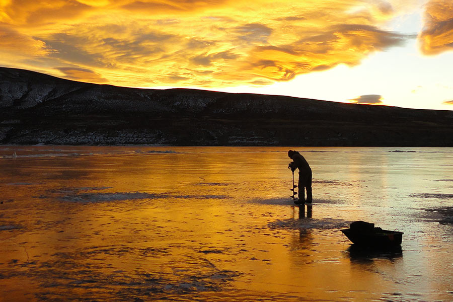 An ice angler drilling a hole with an auger at Flaming Gorge Reservoir at sunrise