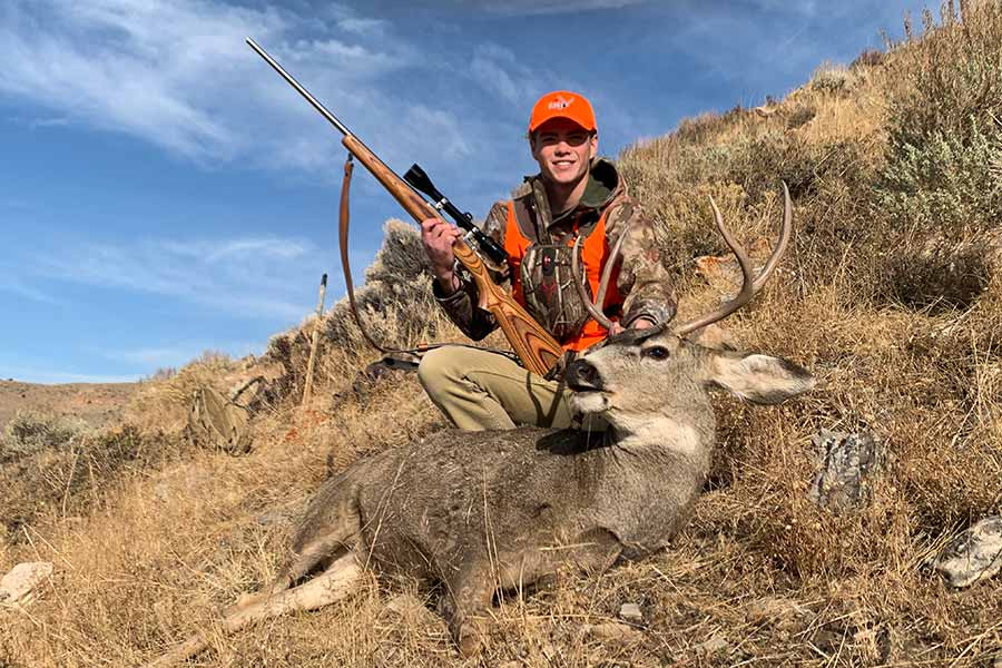 Hunter dressed in hunter orange, holding a rifle and a scope, posing over a harvested deer