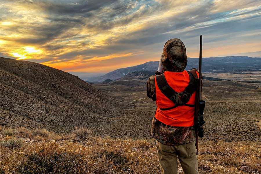 Hunter carrying a rifle and scope looking over a wide valley at sunset